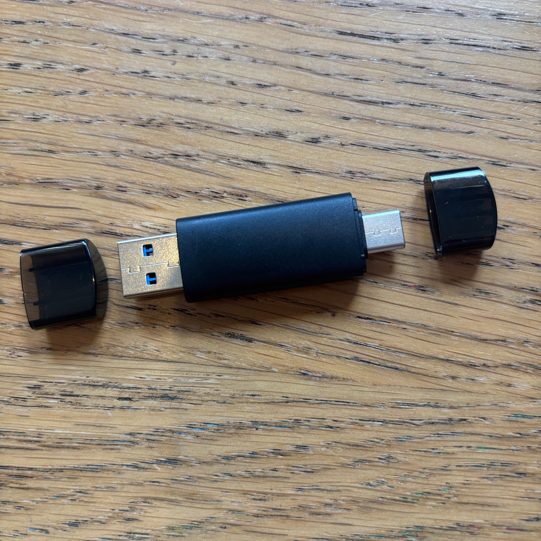 A black USB-C dongle with a USB-A connector exposed, placed on a wooden surface, with its black cap removed and placed to both sides.